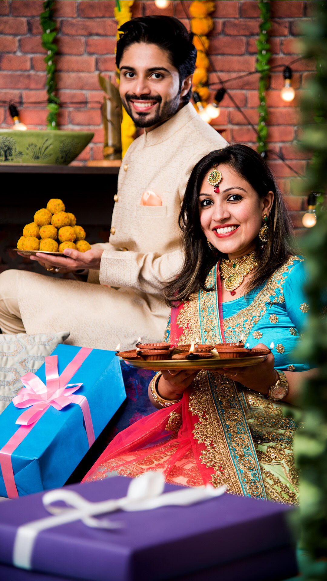groom holding thali of laddos and bride holding thali of diyas with putple gift box near